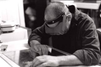 James Munce is sketching a lithograph template with his metal safety googles with his pack of cigarettes nearby. The second view of photographing the subject.