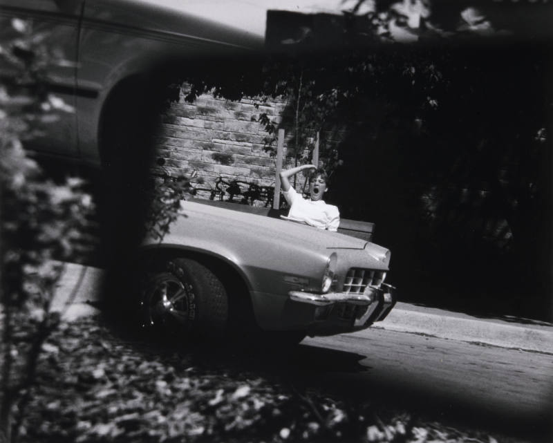 Title unknown (woman yawning on bench behind car)